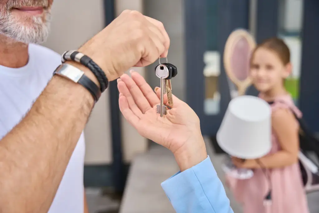 Parent handing house keys to an adult child outside a home while a grandchild waits nearby