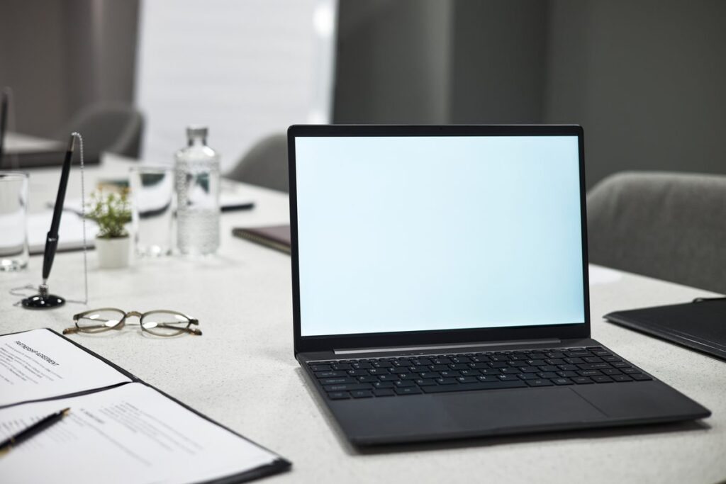 Open laptop on a conference table beside a printed legal agreement, reading glasses and a pen