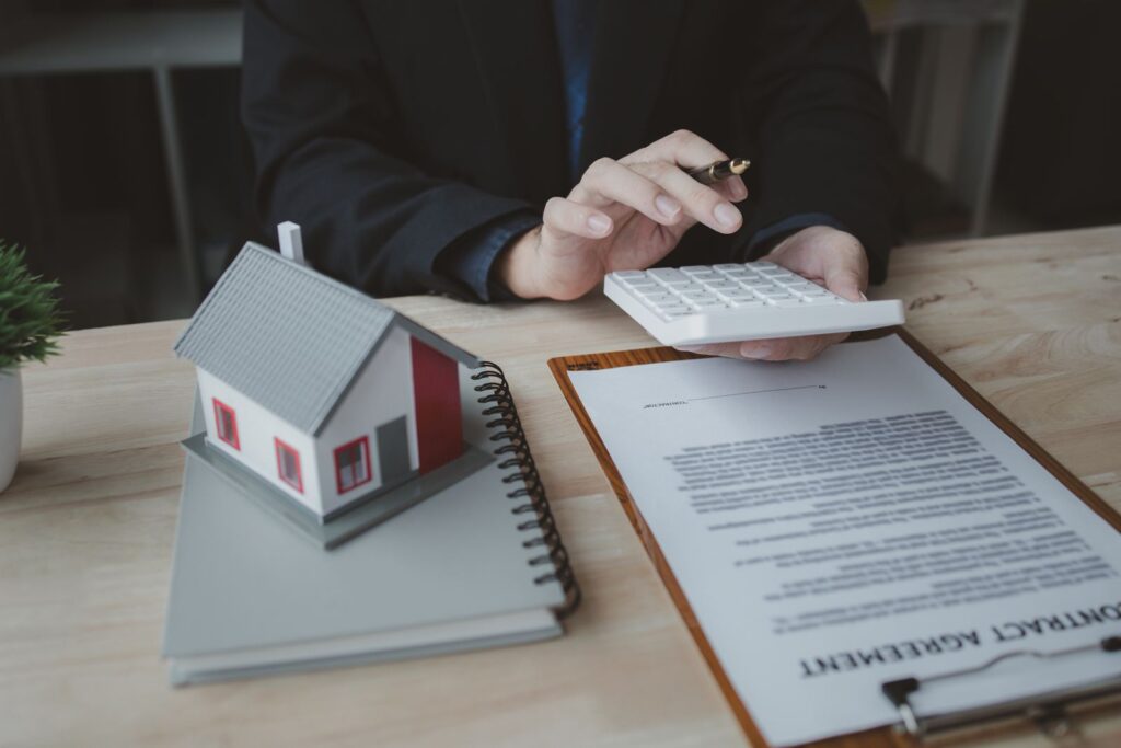 Small model house on a notebook beside a person using a calculator over a contract on a clipboard
