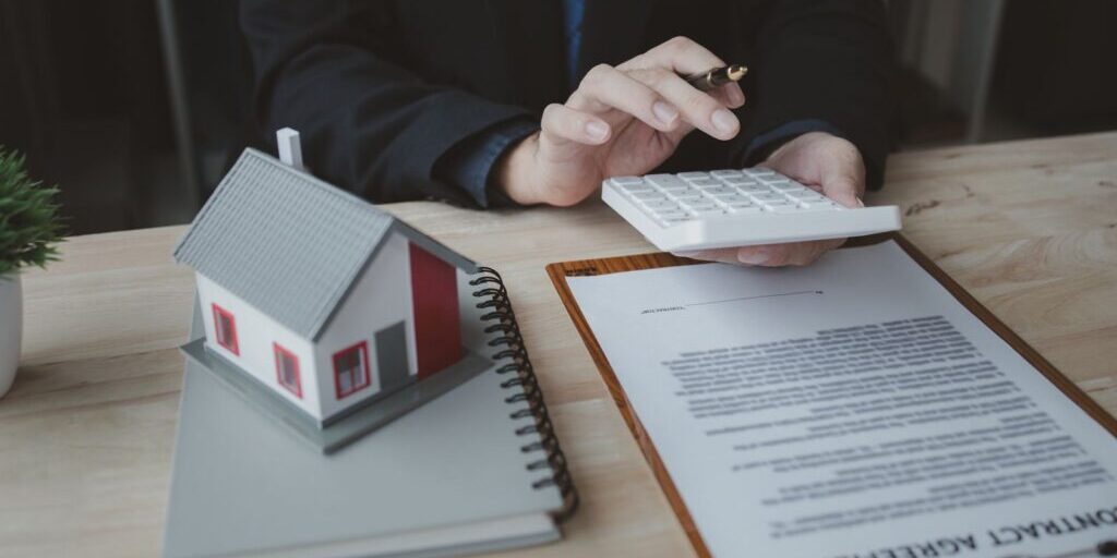 Small model house on a notebook beside a person using a calculator over a contract on a clipboard