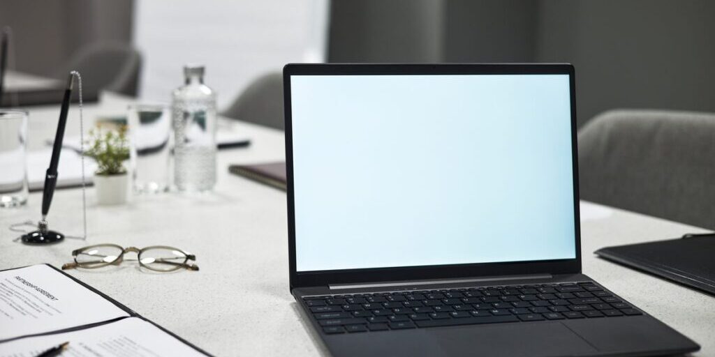 Open laptop on a conference table beside a printed legal agreement, reading glasses and a pen